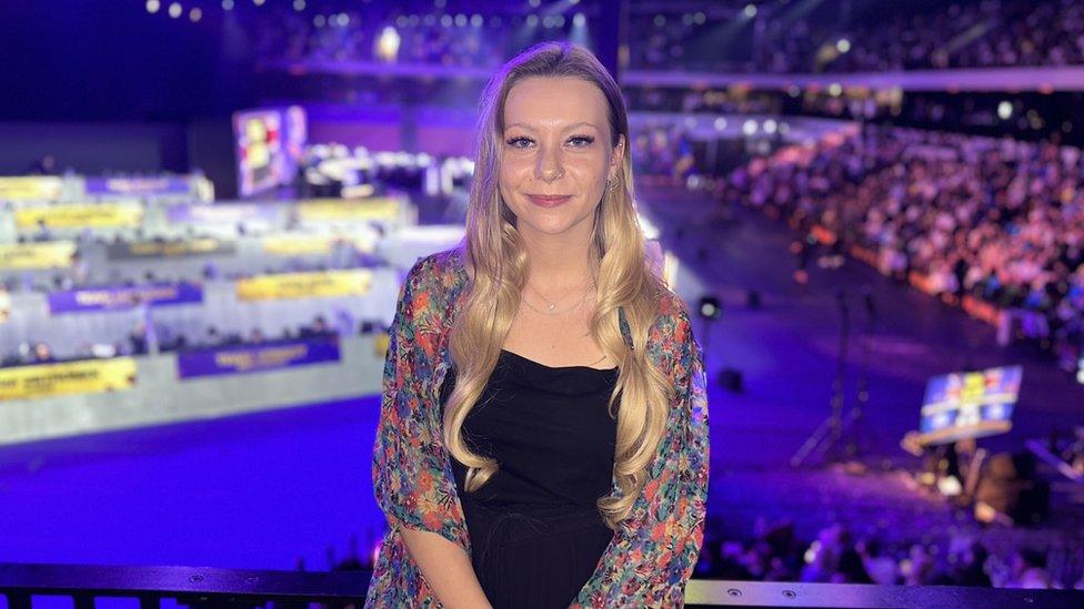 Kels stood smiling for the camera with the arena bathed in the purple glow of stage lights behind her in the background. She has long blonde hair worn down and a ringed piercing in her nose and right ear. She is wearing a black top with a multicoloured flowery patterned shirt over the top of it.