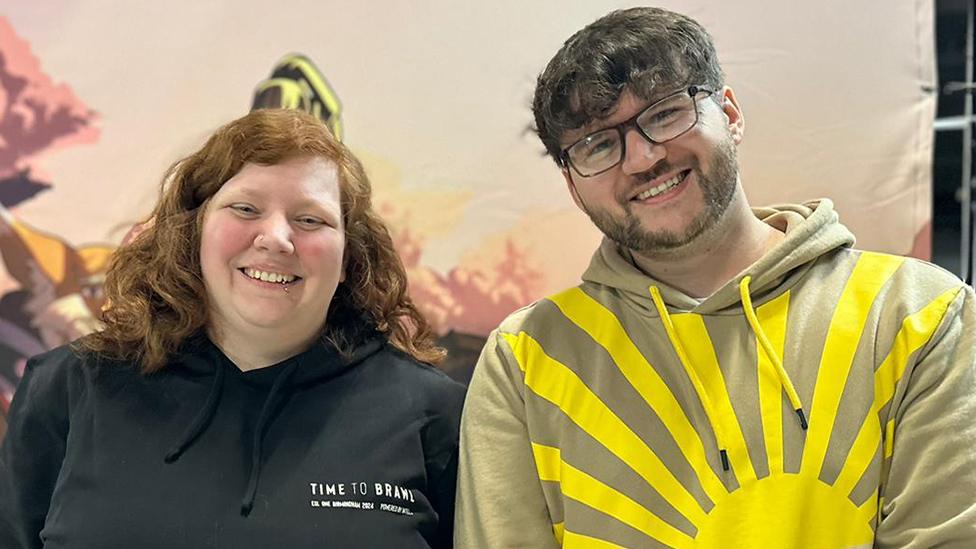 A young woman and a young man stand next to each other, smiling. The woman has curly red hair and wears a black hoodie with "Time to Brawl" written in white block letters on the left chest. The man wears a beige hoodie with a bright yellow sun design - a circle in the lower corner with thick, straight lines representing sunbeams creating a pattern across the garment. He's wearing black-rimmed glasses and leaning slightly towards his friend, who is slightly shorter.