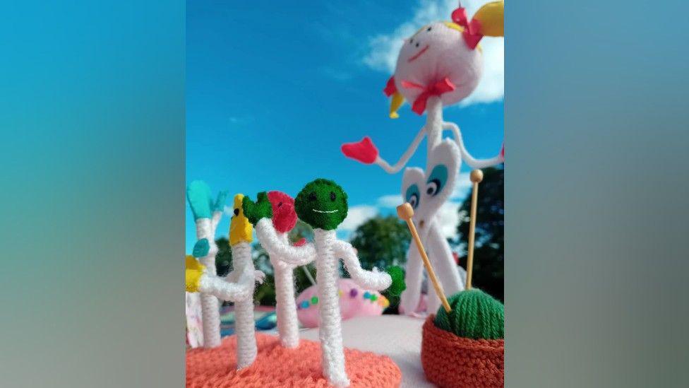 A close-up of the post box topper, showing Polly the pin in the background, with several smaller pin characters with coloured heads. Next to them is a knitted ball or yarn with mini knitting needles stuck in it.