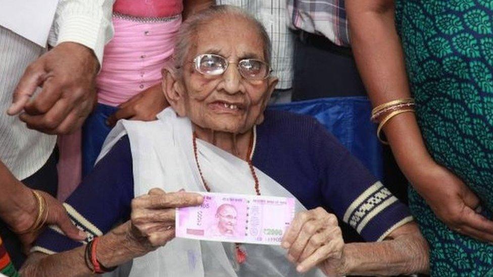 Indian Prime Minister, Narendra Modi"s mother Heera Ba Modi (C) holds up a 2,000 rupee note after exchanging money at a bank in Rysan village, near Gandhinagar, some 30 kms from Ahmedabad on November 15, 2016.