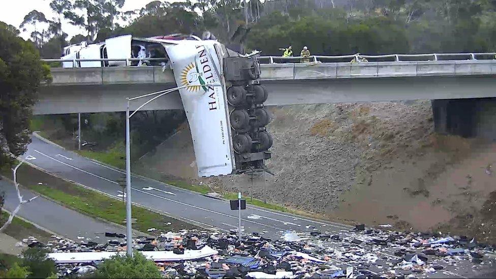 A truck hangs off an overpass bridge with its contents spilled across the motorway below