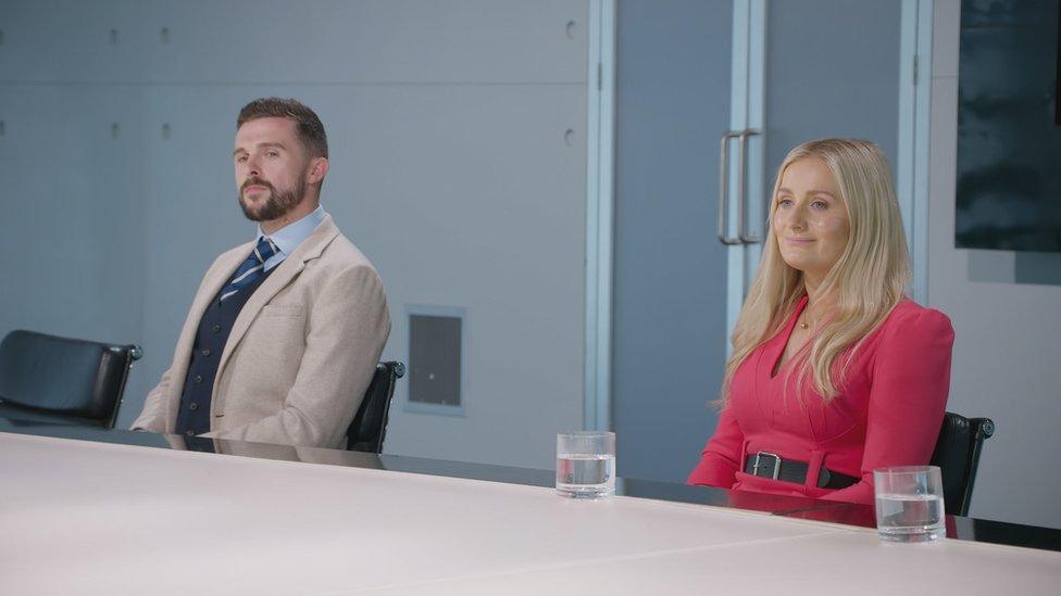 A blonde woman in a pink pantsuit sits at a large table, a glass of water in front of her. To her right a man in a cream jacket, striped tie and blue shirt sits, looking intently at an unseen speaker. The room they're in is quite clinical in appearance, with bare walls and a glass double-door exit.