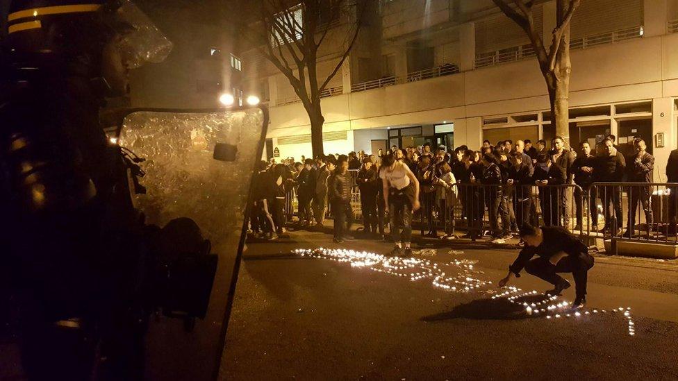 Youths use candles to write the word "Violence" in the road in front of a line of riot police outside a police station in the 19th arrondissement (district) of Paris late on 27 March 2017, during clashes in the wake of the death of a Chinese national during a police intervention