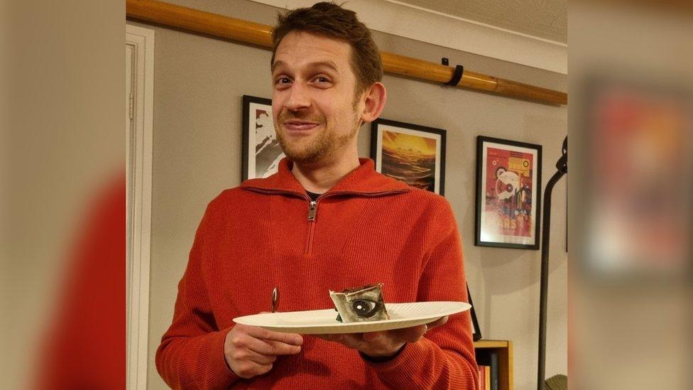 Chris Smowton wearing a red hoodie and holding a slice of cake with a dog's eye on