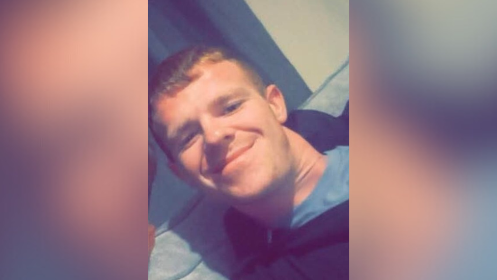 Jamie Mitchell pictured with short light hair. He is wearing a dark hooded jacket and a grey top. He is sitting on a grey sofa.