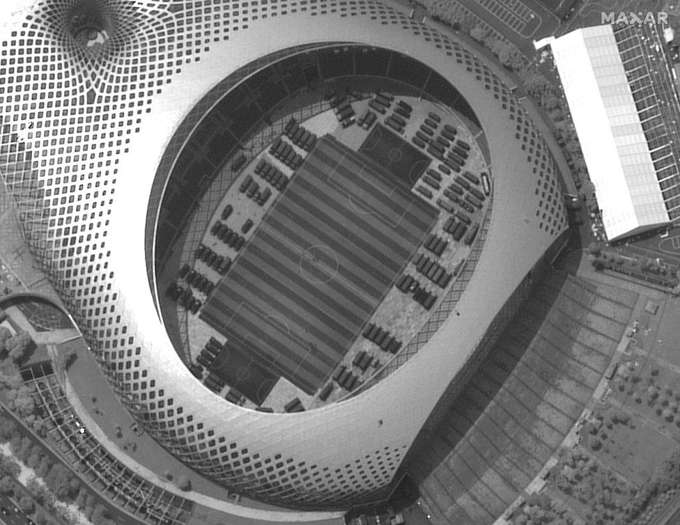 An aerial photograph shows dozens of trucks parked in the interior of a sports stadium, seen through its open roof.