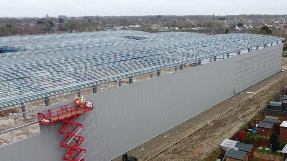 Large metal warehouse building shell, with construction work underway, close to a row of houses