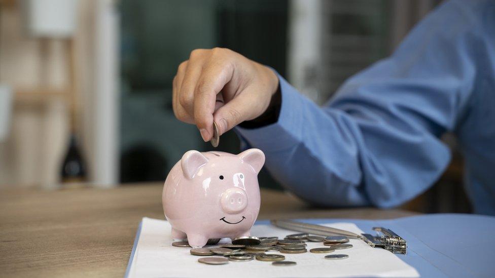 A hand putting money into a piggy bank, with lots of coins scattered around it on a table