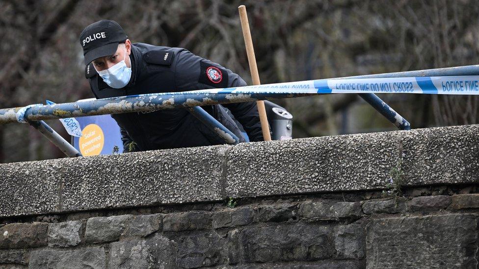 A police officer studies ground looking over a wall while holding a stick