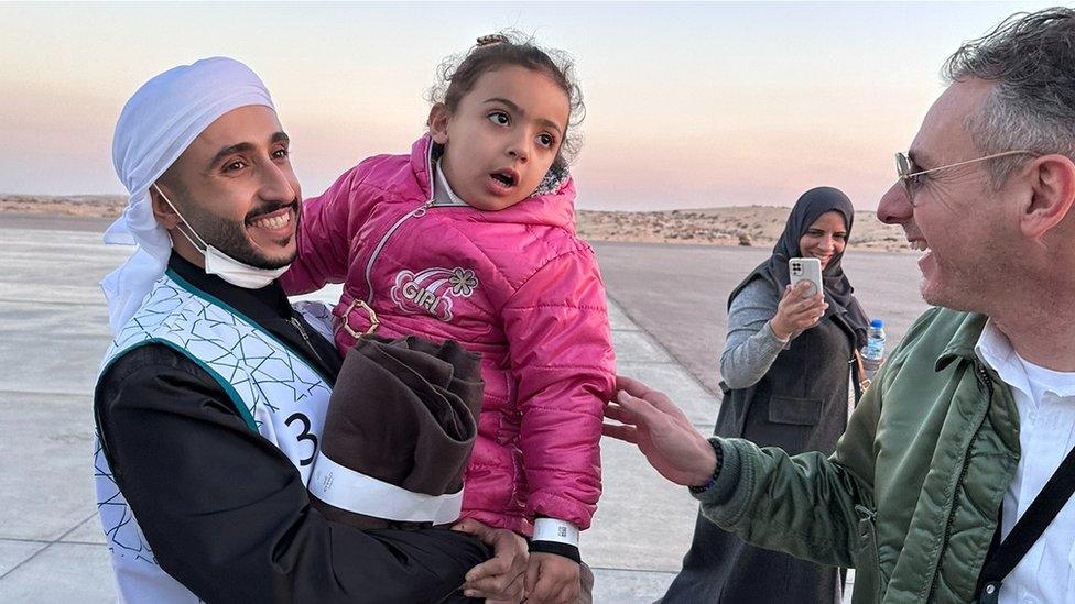 An aid worker is carrying a small child across the tarmac