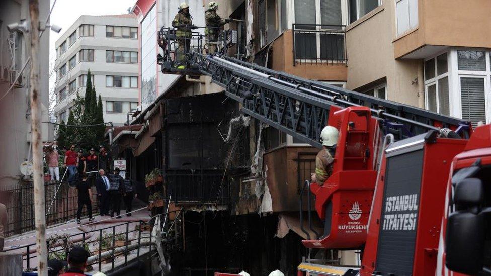 Two firefighter stand on the cherry picker of a fire engine, working at a window. Below them, the building is charred