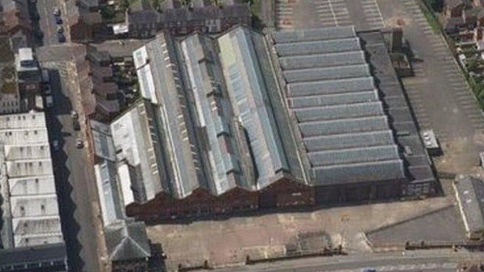 Aerial view showing a line of bus garages with grey roofs, surrounded by houses and trees