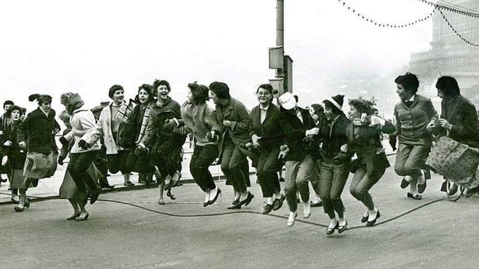 Black and white photo of people stood in a line skipping with a rope