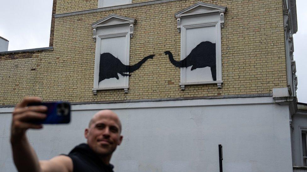 A man takes a selfie in front of a Banksy artwork in London. The artwork shows two elephants painted over windows, their trunks reaching towards each other