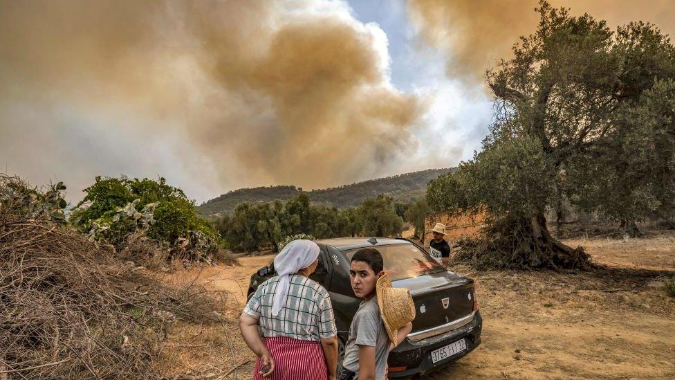 Boy and woman standing outside with smoke behind them and arid land.