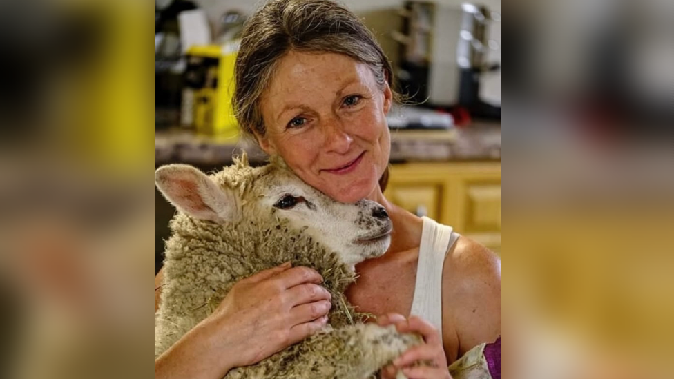 A woman with tied-back grey-brown hair, wearing a white vest top and smiling at the camera. She is caressing a fluffy sheep.