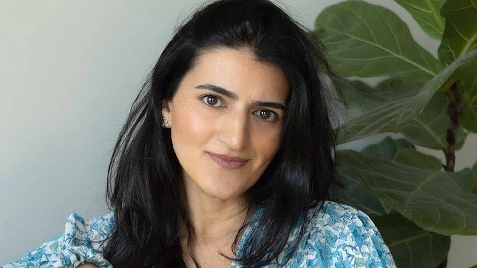 Anoli Mehta wearing a blue and white floral shirt. Sitting in front of a green wall with leaf wallpaper.