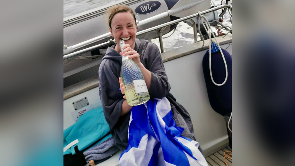 Mairi smiling in the back of a boat, draped in a Scottish flag and clutching a bottle of prosecco