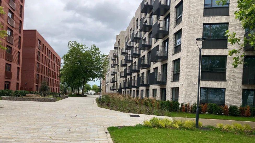 The Athletes Village which has stone pavements, and blocks of flats, trees and grassy verges