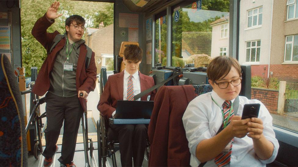 'Mike' (Jack Carroll), 'Sonny' (Zak Ford-Williams), 'Dan' (Reuben Reuter) on the mobility bus, Mike is stood up with his hand raised. Sonny and Dan are both sat down. They're all wearing school uniforms