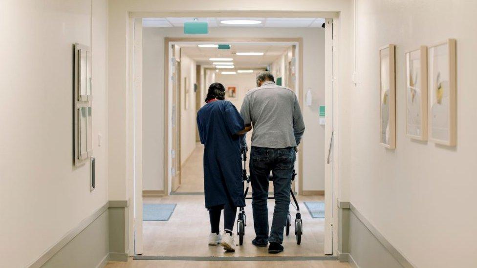 A woman helping a man walk along a corridor