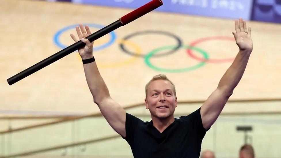 Chris Hoy, wearing a black t-shirt, holds a baton in the air during a ceremony at a velodrome in Paris. Behind him is the velodrome with the five Olympic rings