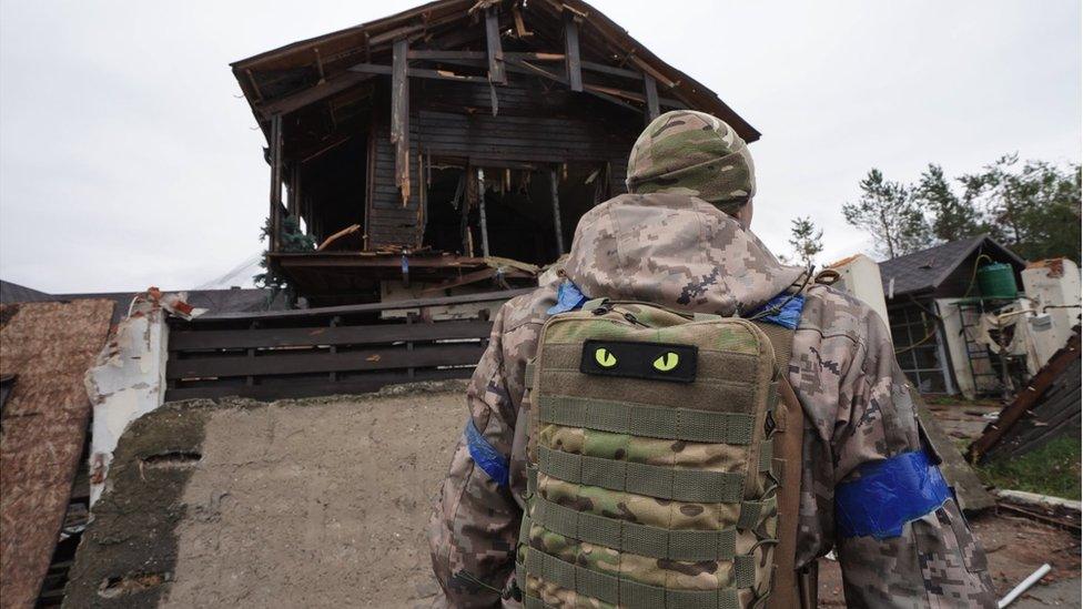 A Ukrainian soldier stands in front of a ruined house