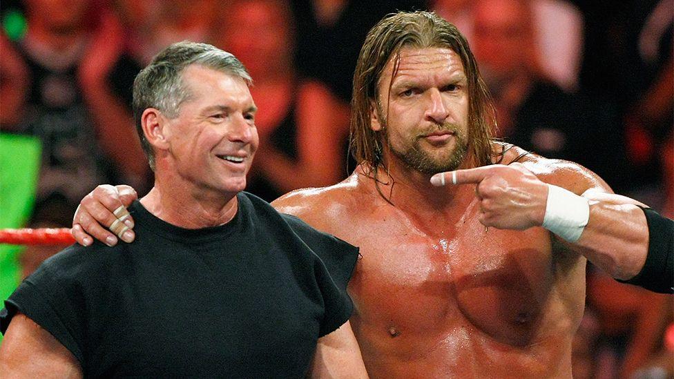 Vince McMahon and Triple, standing in the ring, with Triple H pointing towards Vince. There is a full crowd behind them.