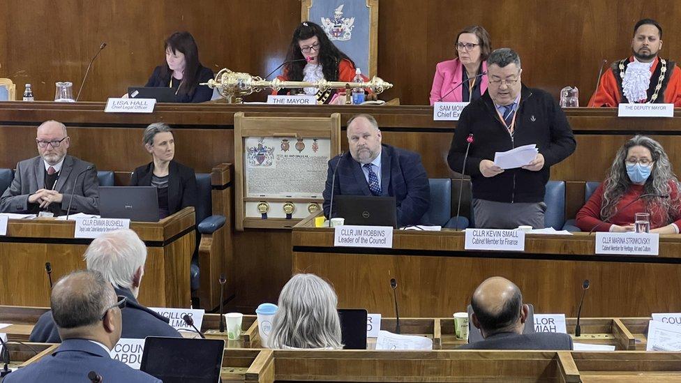 A council meeting with councillors sat at wooden benches.