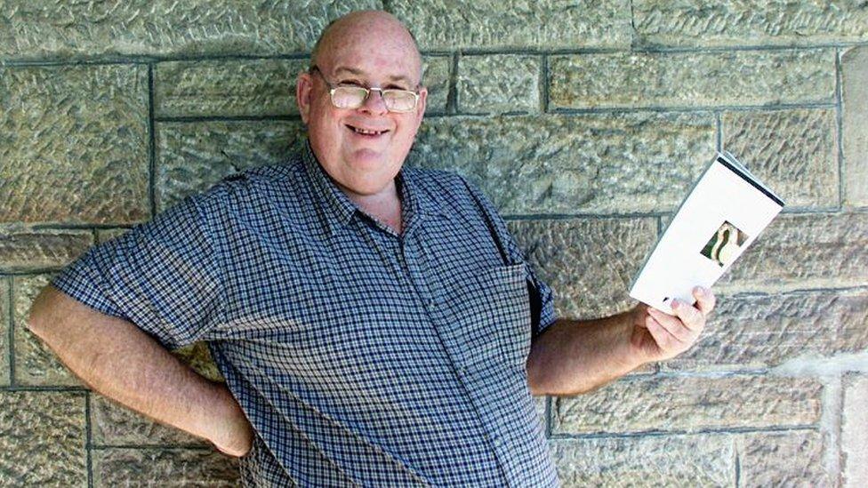 Les Murray stood outside, wearing glasses, smiling, holding an book, looking at the camera