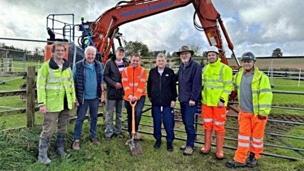 A group of eight councillors and contractors are photographed in a field with an orange digger behind them. Those photographed are seen in high viz clothing.