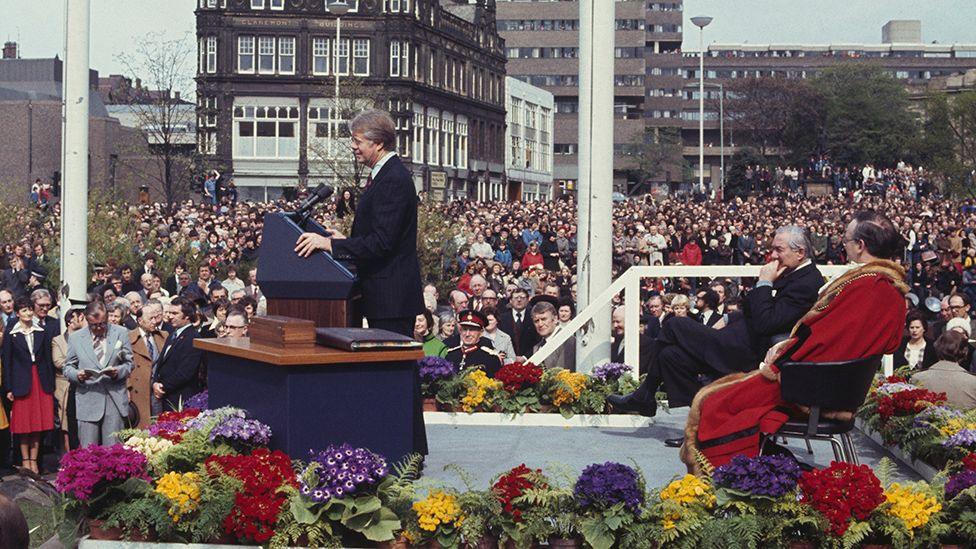 Former president Jimmy Carter greeting a crowd in Newcastle in 1977. There are flowers around the podium, on which sits the Lord Mayor in red robes, and Prime Minister James Callaghan.