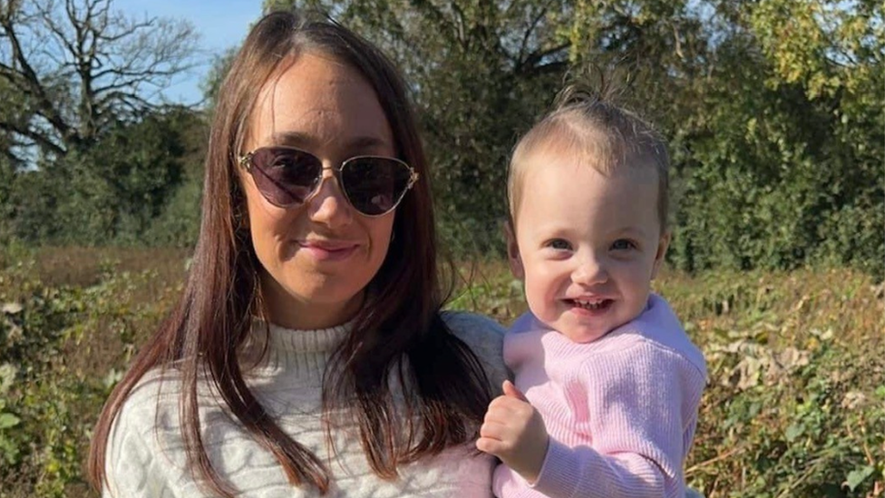 A mother in sunglasses holding her toddler and both are smiling at the camera