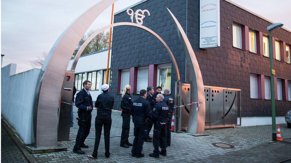 Eight people, most of them visibly in police uniforms, stand outside a building where police tape is visible