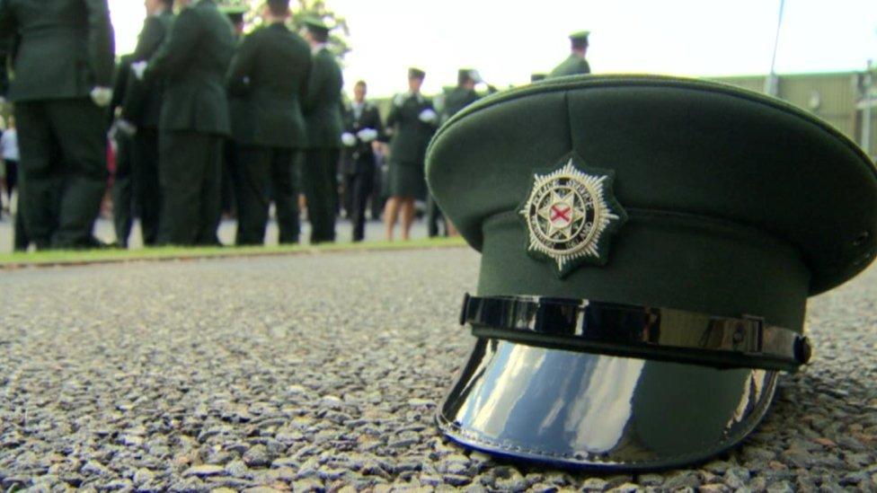 A PSNI cap sits on the ground, in the background officers are standing casually chatting at their passing out parade