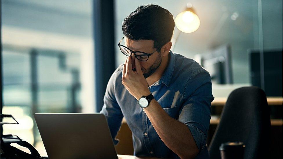 A stressed man pinches the bridge of his nose in frustration in front of his laptop in this photo illustration