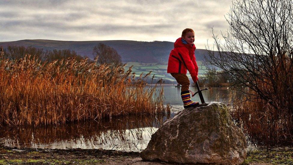 A photo of a child holding the Excalibur sword in the stone by the lake.