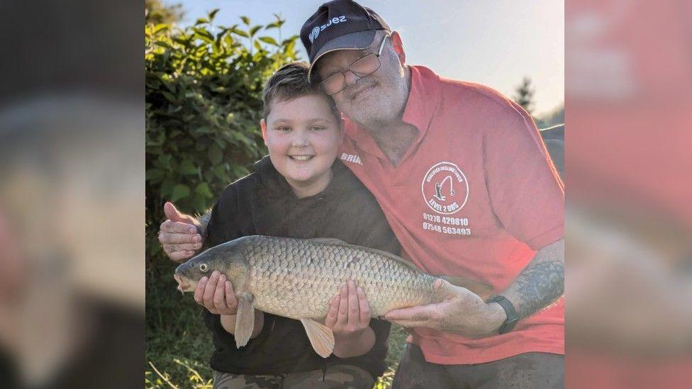 Mr Barker sitting with a boy holding a fish. Mr Barker is wearing a baseball cap and salmon shaded top. The boy has brown hair and is wearing a black T-shirt.