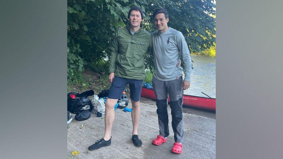 Charlie Goodwin, wearing a green top and blue shorts, and Hector Bevan, wearing a grey top, grey trousers and red shoes in front of a canoe