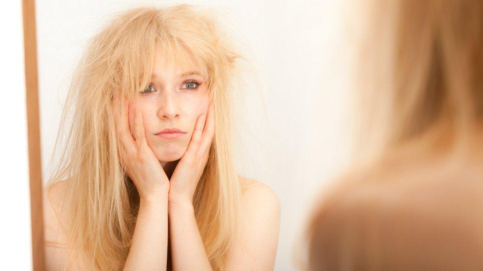 A young blonde woman, head in hands, fed up with her hair as she looks into the mirror