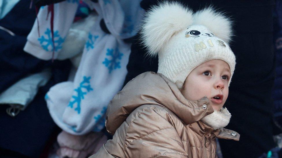 A small child walks around a pile of clothes at a makeshift camp