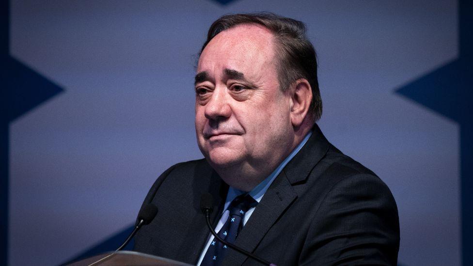 A man with dark hair and dark, bushy eyebrows sits at a 45 degree angle to the camera, with a microphone in front of him. Behind him is a saltire background. He is wearing a dark suit, blue shirt and dark blue tie with saltires on it. 