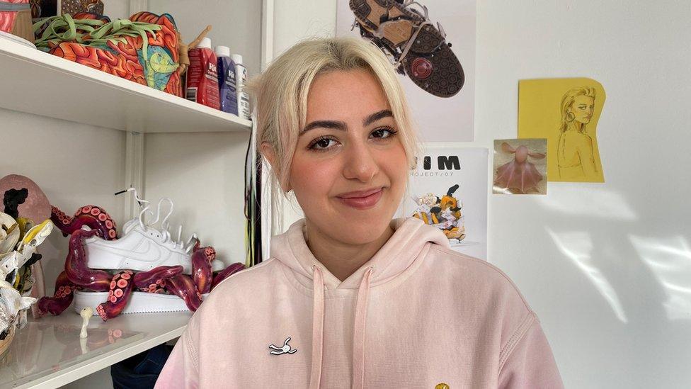 Melicka has blonde hair and is wearing a pink hoodie. She is sitting in front of a shelf with her artwork