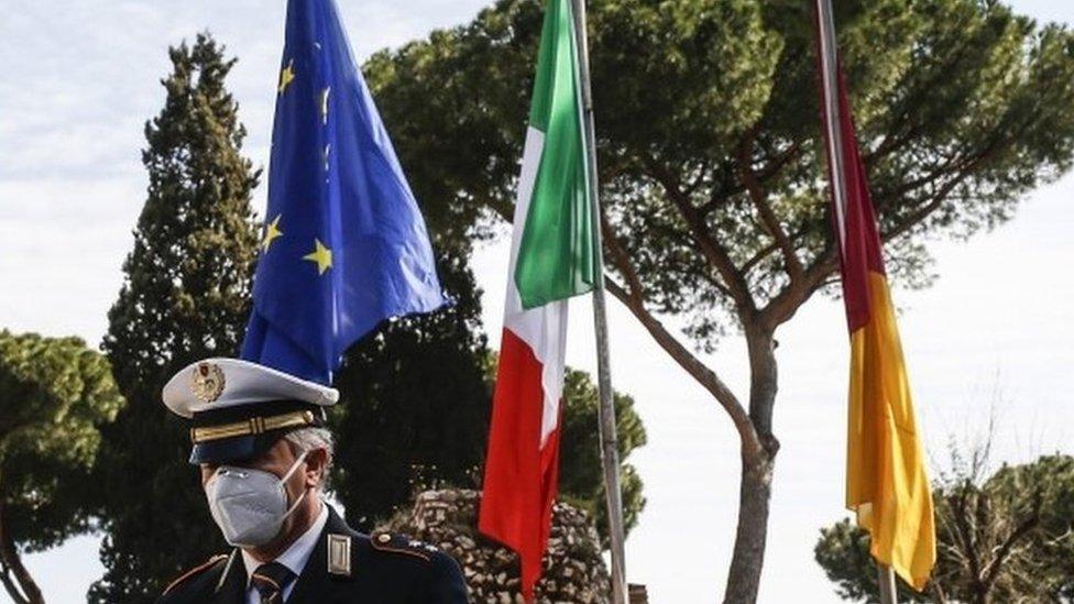 An Italian police officer stands next to EU, Italian and German flags in Rome. Photo: 31 March 2020