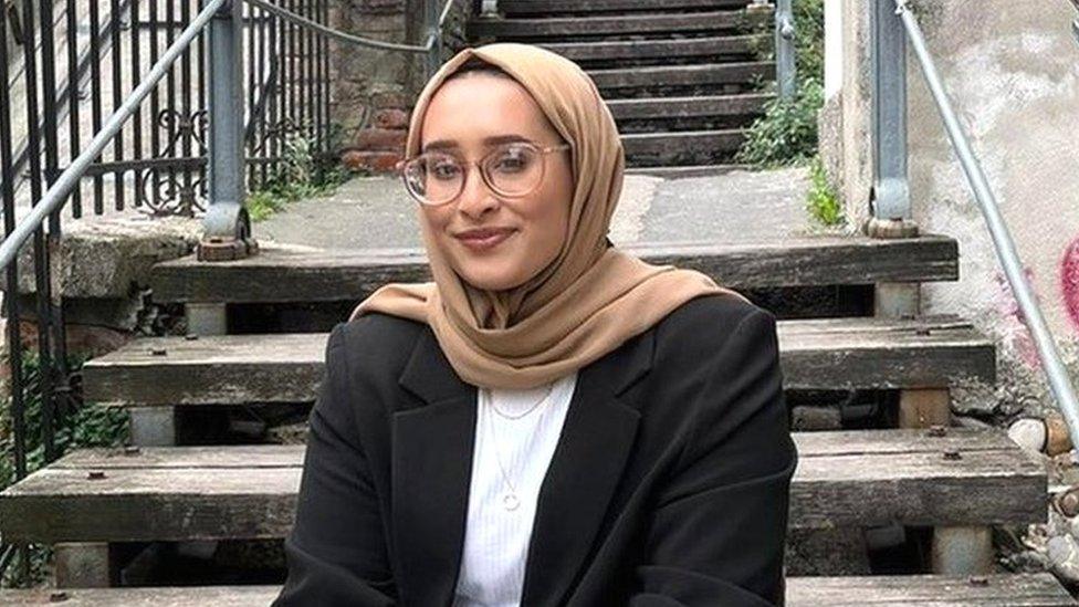 Female wearing beige headscarf, white top and a black blazer, sitting outside on steps.