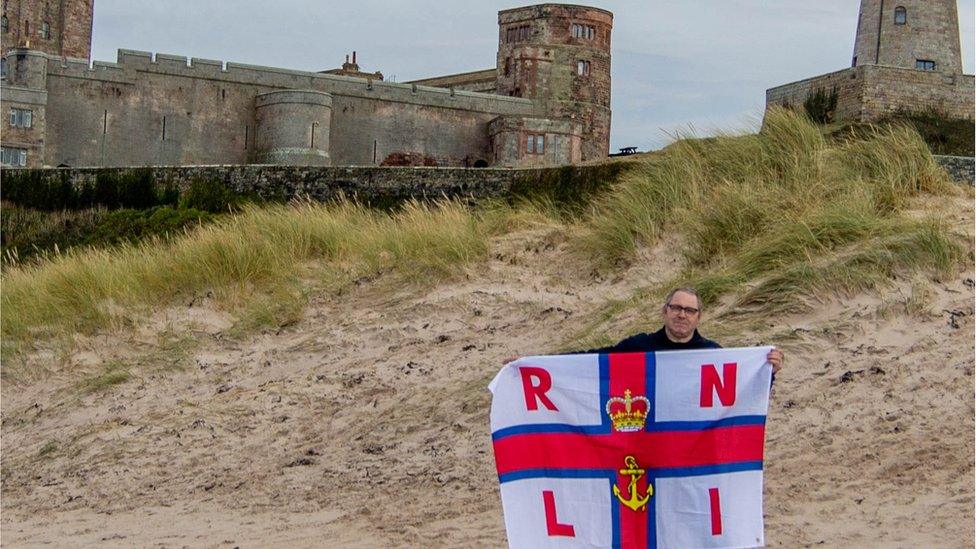 Chris Mason holds RNLI flag beside Bamburgh Castle