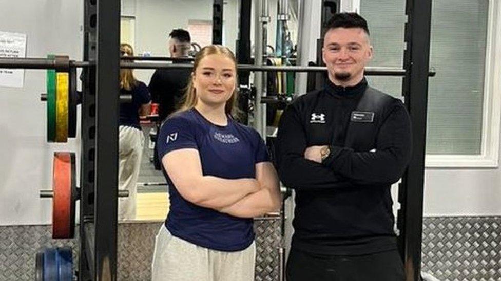 Phoebe Pothecary standing in front of a weight rack at the gym with her arms folded, beside another man