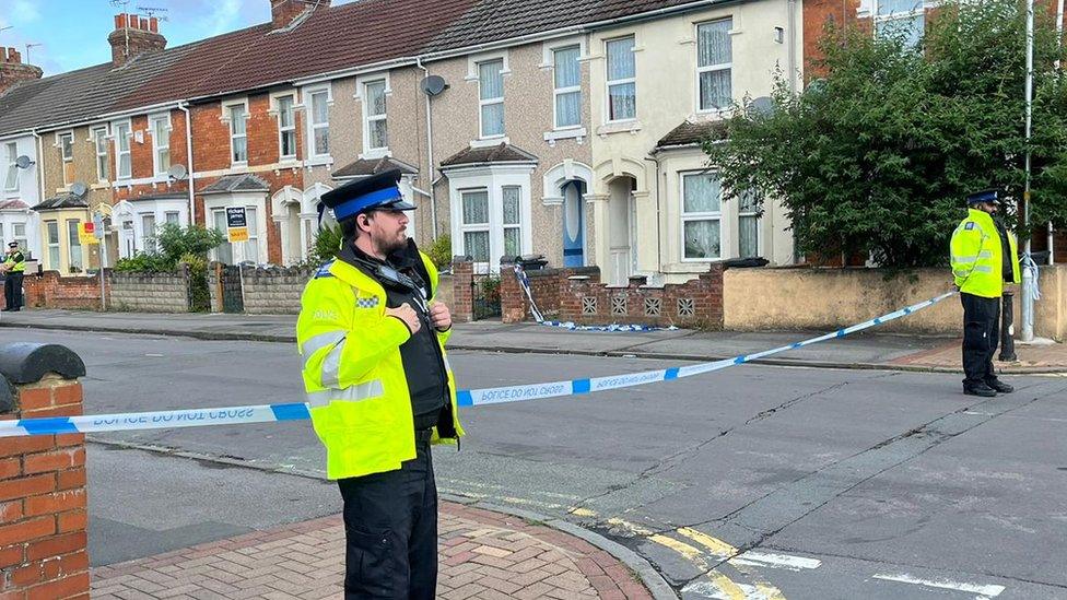 Two police officers stand next to a road blocked off with tape