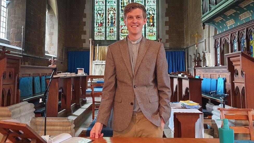 Man with short light brown hair and brown jacket with clerical collar stands in a church