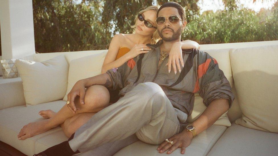 A young white woman and a slightly older black man sit on an expensive-looking sofa on a white marble veranda. They appear to be in a sunny country. Both wear sunglasses and she's sitting behind him, leaning on his shoulder. She's wearing a orange tube top and a skirt, her bare feet are curled up and resting on the edge of the couch. He's wearing a dark grey tracksuit top with black and red accents which looks retro. He's wearing trousers in a matching shade and resting one arm on the woman's knees.